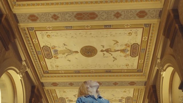 View looking up at a woman notices the ceiling art among golden archways, 23.98 fps.