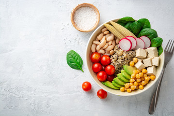 Healthy vegan buddha bowl with spinach, tomato, chickpea, quinoa, radish, beans, avocado, mini corn and tofu cheese on concrete background. Top view. Copy space.