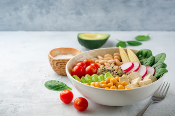 Healthy vegan buddha bowl with spinach, tomato, chickpea, quinoa, radish, beans, avocado, mini corn and tofu cheese on concrete background. Selective focus.