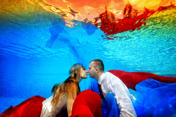 Fabulous wedding underwater. The bride and groom swim and kiss underwater in the pool at sunset surrounded by red and blue fabric. Portrait. Concept. Underwater photography. Horizontal view