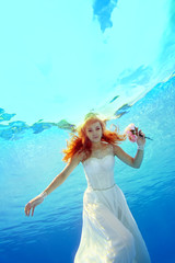 Fototapeta na wymiar A charming bride with red hair swims and poses for the camera underwater in the pool in a white dress with flowers in her hand on a Sunny day. Portrait. Bottom view. Underwater photography