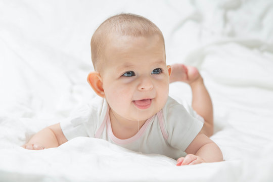 Increased salivation in the infant during teething. A baby girl in white clothes is lying on a white bed, smiling, drooling. Salivary glands activation