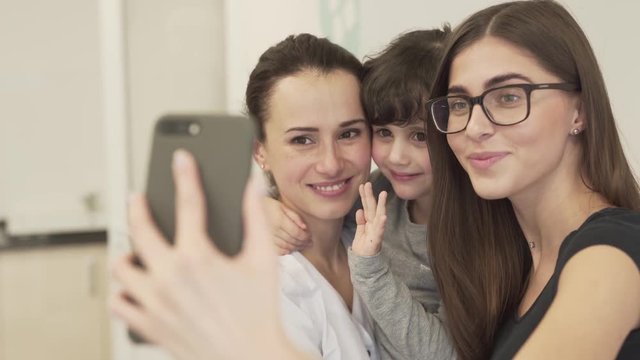 The dentist holds a baby girl and she hugs the doctor. The elder sister stands next to them and holds a phone in order to take a selfie. They all smile and pose for the picture. Background is blurred.