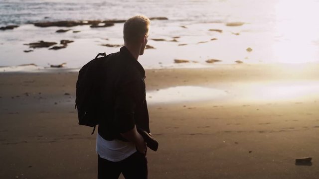 Man walks along black sand beach, does flip against golden light of sunset, slow motion, lens flare