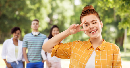 gesture and people concept - smiling red haired teenage girl in checkered shirt showing peace hand sign over group of friends at summer park background