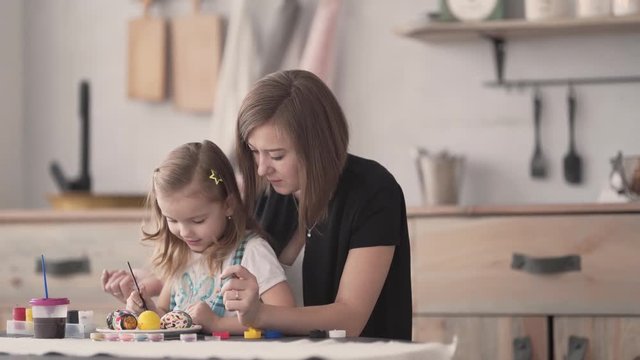 Daughter sitting on her mum's lap at table and painting the Easter eggs with gouache ushing paintbrush. Mum's helping daughter draw and paint. Happy family concept