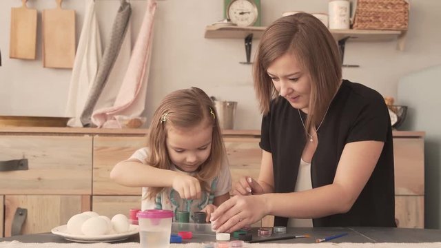 Mother and daughter sit in workshop and are about to paint. They try to open little dried paint cans. Child gets some of black paint on her fingers, shows to her mum. Mother smiles and shakes her head