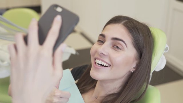 Close-up of a young cute woman taking selfies of her magnificent smile in the dental office.