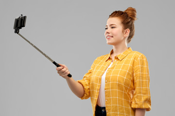technology and people concept - smiling red haired teenage girl in checkered shirt taking picture by smartphone on selfie stick over grey background