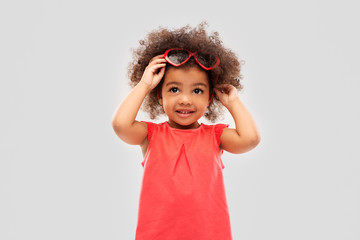 childhood, valentine's day and summer concept - happy little african american girl with heart shaped sunglasses over grey background