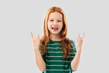 childhood, gesture and people concept - smiling red haired girl in green striped shirt showing rock hand sign over grey background