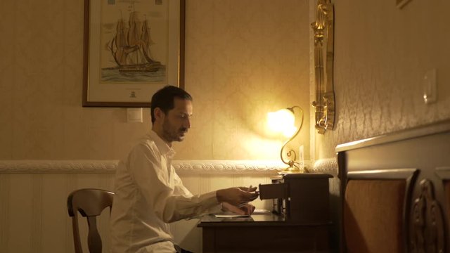 Man in white shirt sitting in a dark room with a table light takes out of a wooden desk drawer his pen and is getting ready to write.