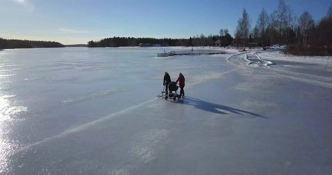 Aerial, young woman with baby stroller and older woman with kick sled walking on frozen lake on a beautiful sunny winter day