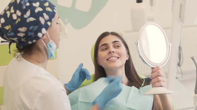 Dentist holds wide and proper consultation about patient's teeth, jaw and bit condition. Girl sits in the chair and holds a mirror to see the full image. Dentist uses mouth mirror to show all flaws.