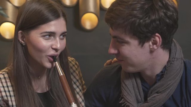 A beautiful girl and a handsome boy sit in the club and enjoy themself. The lady smokes hookah and smiles. The gentleman hugs and contemplate her. They talk. The background is blurred