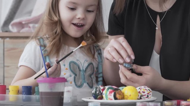 Small blond girl holding a paintbrush in her right hand and trying to paint Oster eggs. Child is smiling a lot and very happy. Mum's helping girl painting with gouache