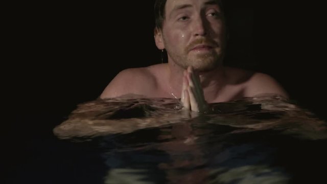 A guy swimming at night emerges from the water and looks around despondent