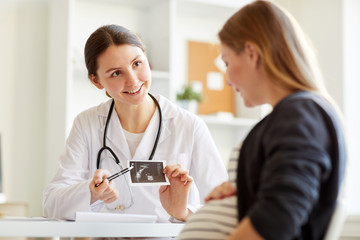 Portrait of smiling onstetrician showing ultrasound scan of baby to pregnant woman visiting doctor, copy space