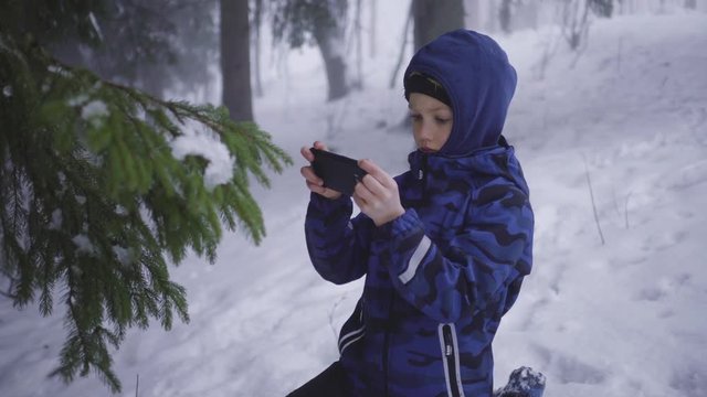 Young boy in dark blue jacket and black pants making photo with black smartphone of spruce branches - snowy mist in winter forest
