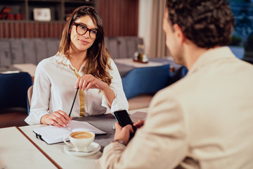 Beautiful Caucasian businesswoman dressed elagent holding agenda and looking at camera. In foreground her colleague sitting at table. Restaurant interior.