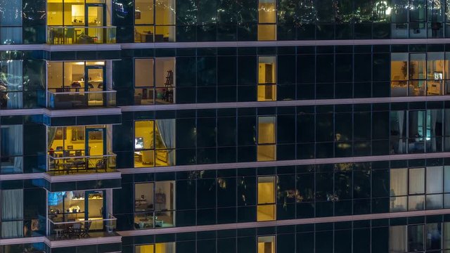 Glowing apartment windows at night in glass skyscraper timelapse. Warm light and motion inside. Lights reflected from surface. Pan right