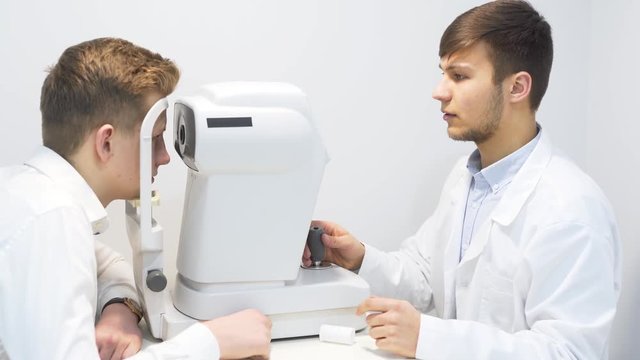 Male Optician Examining Male Patient Eyes While Using Equipment During Checkup Panning Left