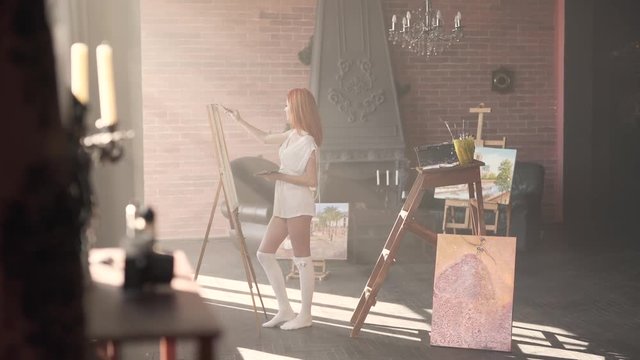 Wonderful young red-haired girl is standing in her workroom in only white transparent shirt and painting. The room is made in a loft style.