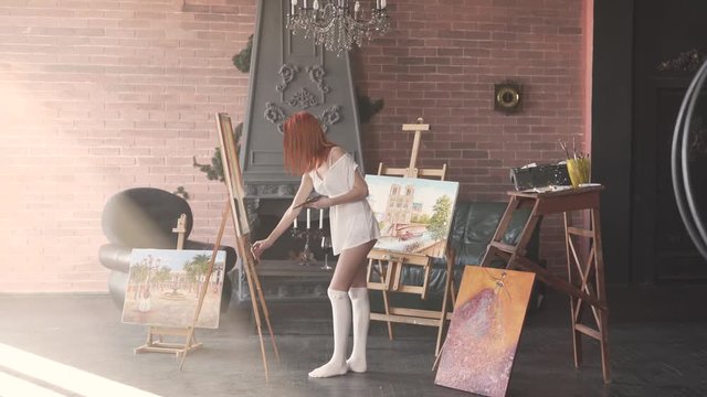 Sexy young red-haired woman is standing in her workroom in only white transparent shirt and painting. The room is made in a loft style.