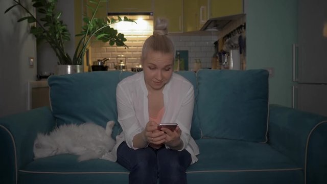 A young woman uses the phone sitting on the couch in the evening with a cat. The girl read the good news on the phone. 4K