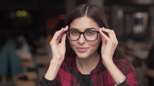 Young Woman Is Looking Directly Into The Camera, Smiling And Putting A Glasses On Her Face.