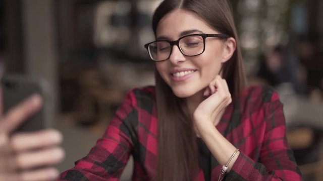 Portrait of a young charming woman who is sitting in a cafe indoors and talking in a video chat with her friend and smiling. She is looking absolutely relaxed and easy.
