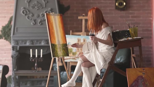 Seductive young red-haired girl is sitting on a chair in front of a picture she is painted holding a glass of red wine and correcting something with a brush and paints.
