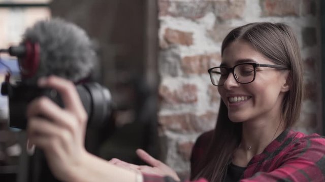 Beautiful young girl in a glasses is sitting in a cafe and making a selfie with a professional photo camera. She is changing her face expression to make a funny photos