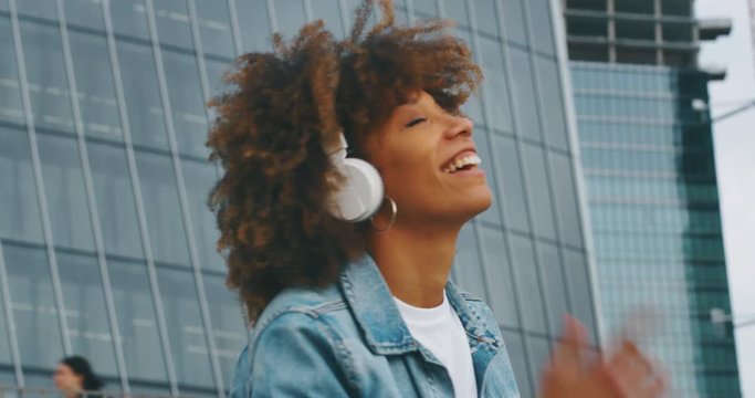 Slow motion of young african woman is having fun to listening to the music with white headphones and dancing in a city center with skyscrapers.