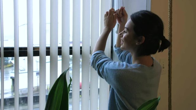 A happy girl opens the blinds to let the light into her apartment.