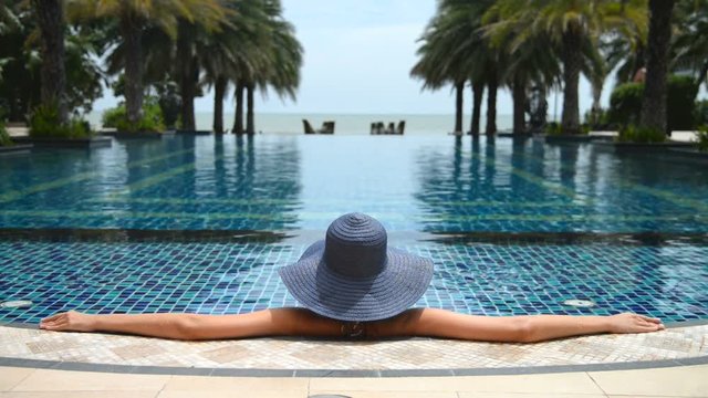 Young woman in a blue straw hat relaxing with arms stretched out along the lege of resort infinity pool looking out into the ocean​ beyond.