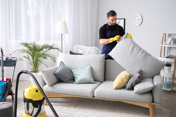 Male janitor cleaning sofa in room