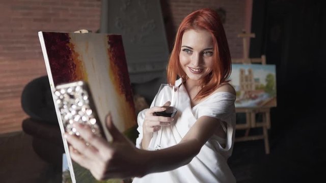 Young beautiful female painter is making a selfie on a phone. She is standing near her new picture, smiling and holding in a hand a glass of wine.