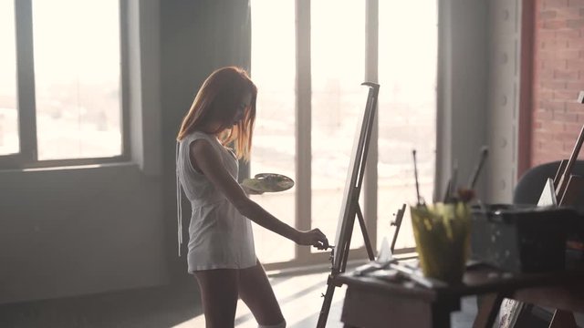 Red haired woman painting on canvas in front of a window