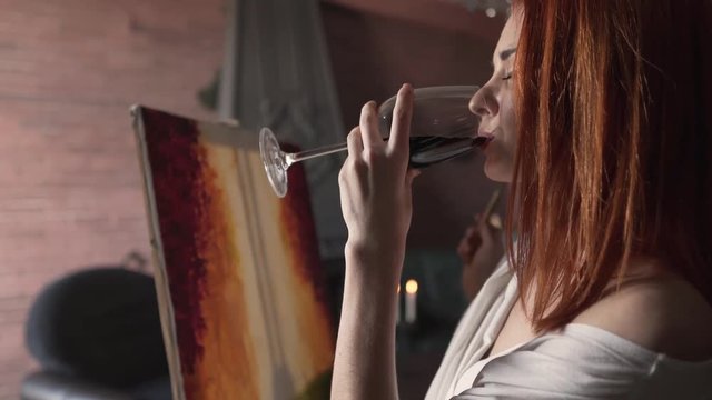 Attractive woman sips from a glass of red wine as she paints.