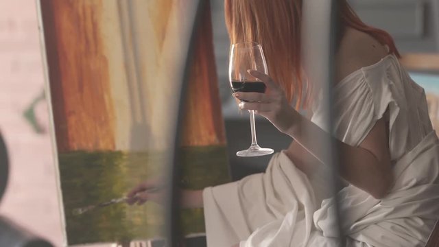 Beautiful artist holds a glass of wine with one hand and uses a paintbrush with the other to work on her colorful painting.