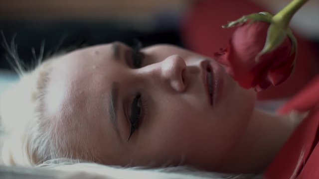 In slow motion, a young woman lies on a bed holding a single red rose and smiles at the camera confidently