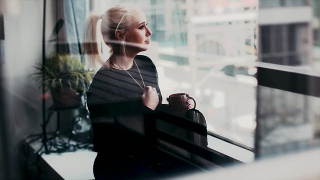 A young blonde woman gazes outside her large city apartment window and plays with her hair
