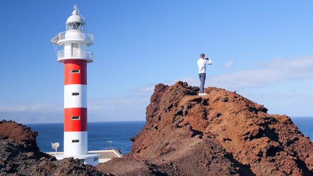 View of a tourist standing next to a lighthouse on a volcanic cliff shooting the landscape. Located at Punta de Teno, Tenerife (Canary Islands). HD cropped edit