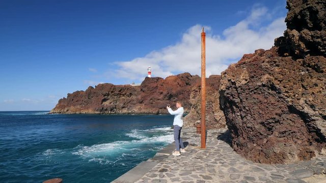 View of a tourist standing at the dock and shooting the volcanic cliffs and the famous lighthouse of Teno in the Isle of Tenerife in slow motion