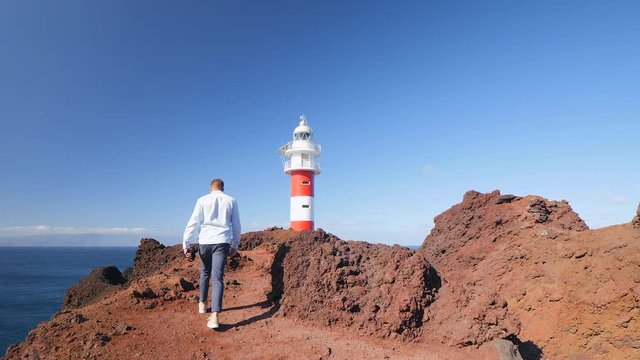 Follow back view shot of a tourist walking at the edge of a volcanic cliff towards the famous lighthouse of Teno in the Isle of Tenerife, then stops and takes a photo in slow motion