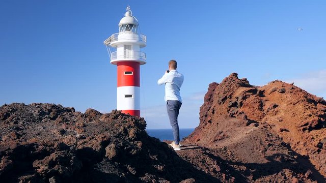 View of a tourist standing on a volcanic cliff and photographing the lighthouse of Teno, Isle of Tenerife . Seagulls are flying and La Gomera island is in the background. HD edit cropped