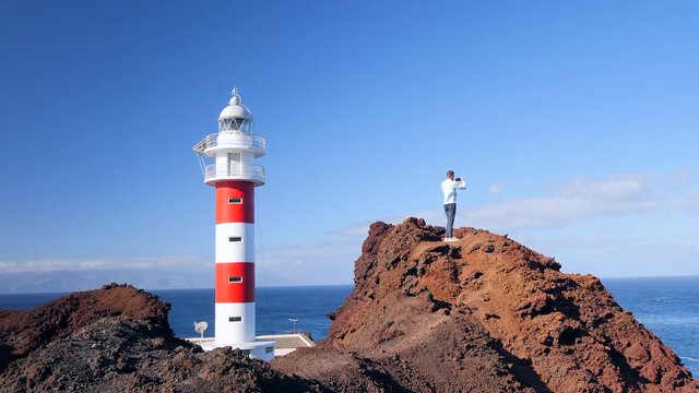 View of a tourist standing next to a lighthouse on a volcanic cliff shooting the landscape. Located at Punta de Teno, Tenerife (Canary Islands)