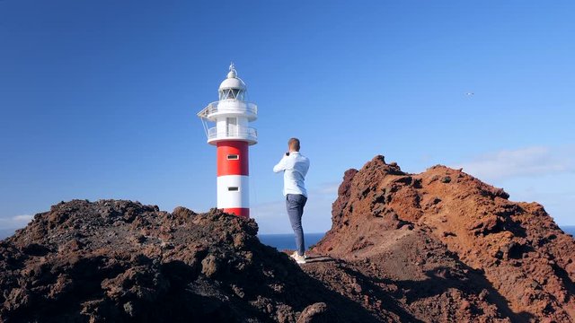 Pedestal camera view of a tourist standing on a volcanic cliff and photographing the lighthouse of Teno, Tenerife (Canary Islands). Seagulls are flying and La Gomera islands is in the background