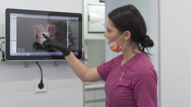 Female Dentist in Her Uniform and Gloves in Modern Dental Clinic Showing Scans Details on Monitor to Her Patient.